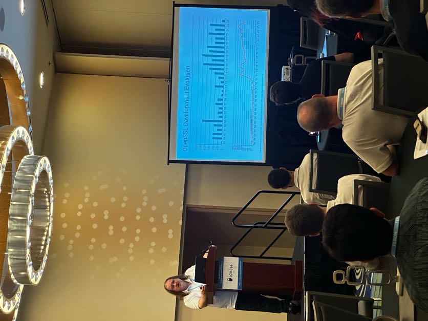 Tim Hudson at the podium with a slide titled OpenSSL Development Evolution showing a bar chart of development activity since 1998.
