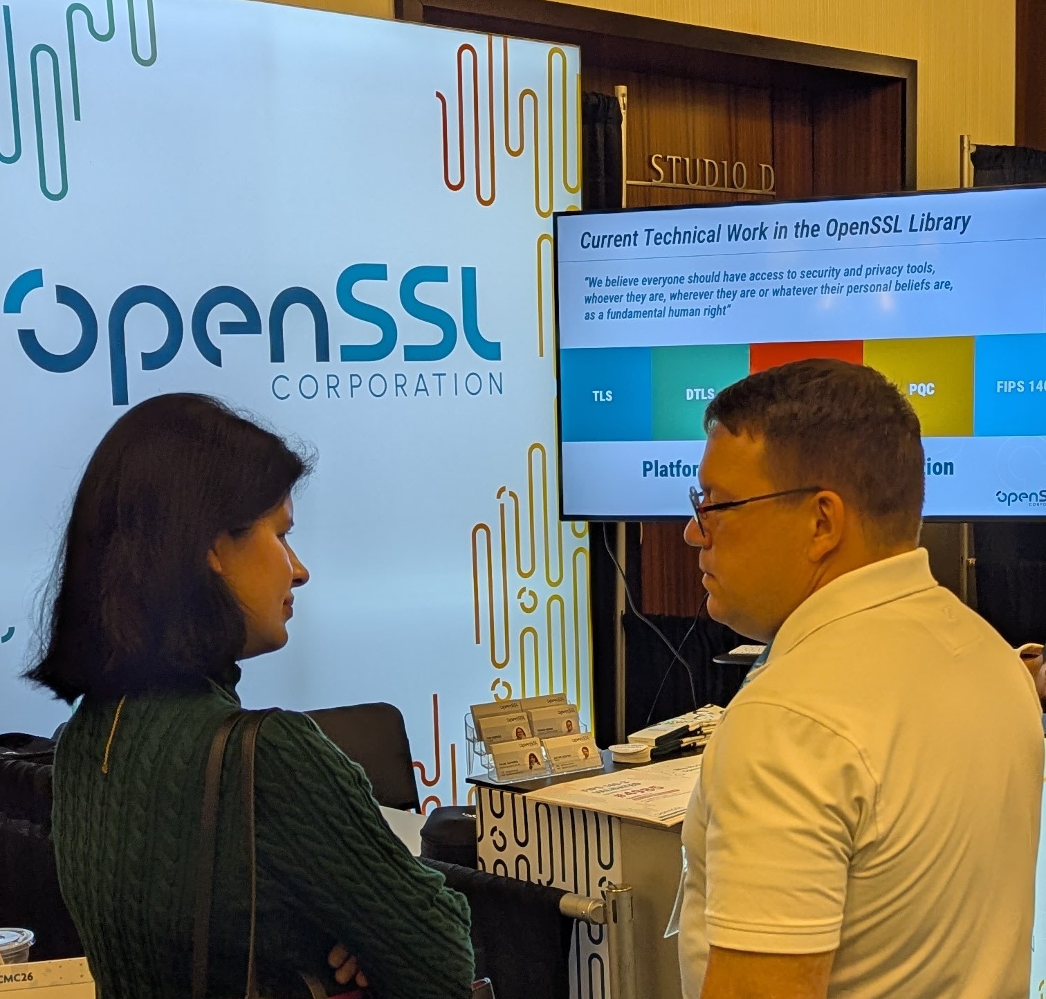 Tomáš at Booth 4, in conversation with an attendee, the OpenSSL Library technical-work slide on the screen behind.
