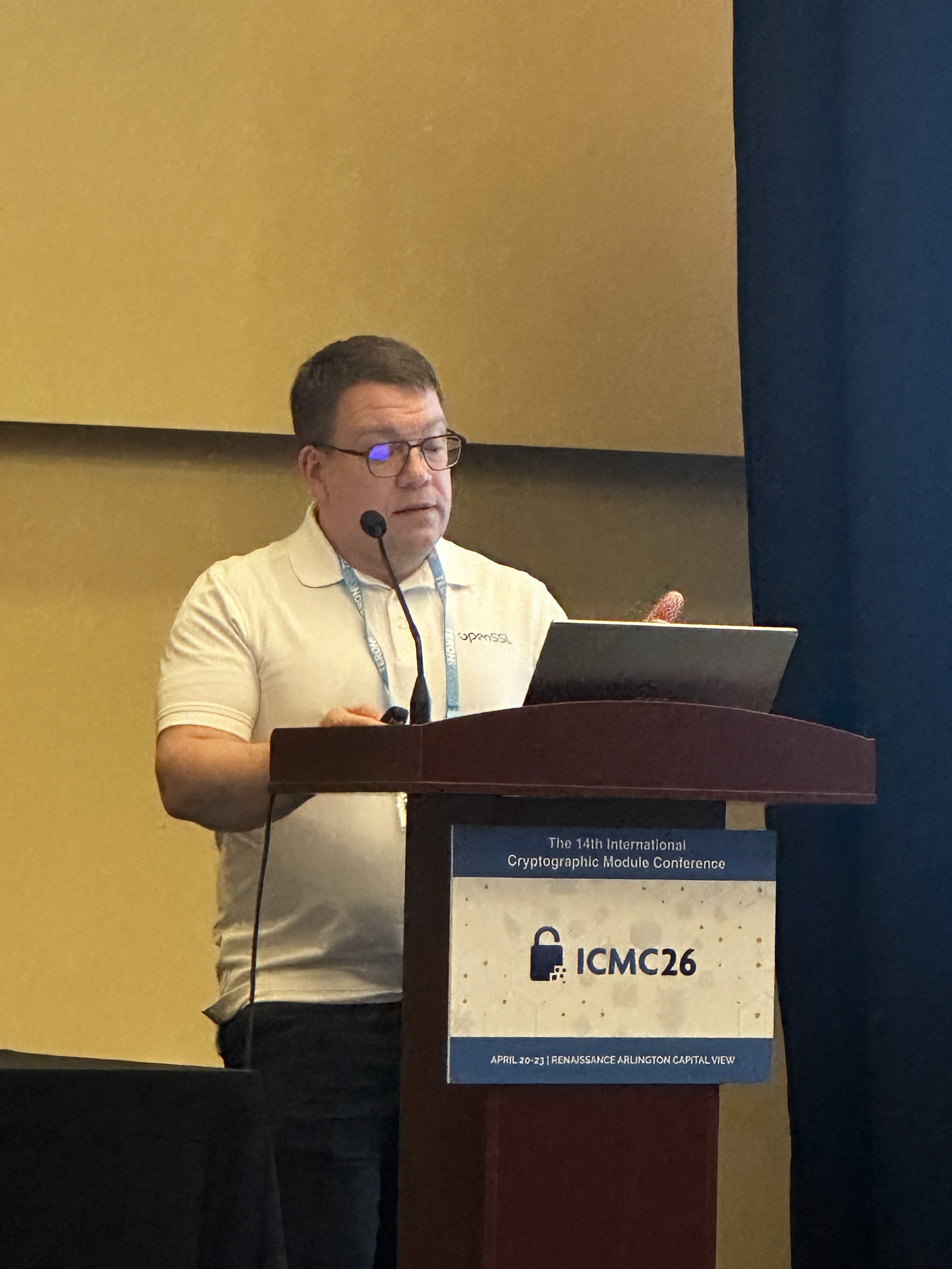 Tomáš Vávra reading from his laptop at the ICMC26 podium, OpenSSL polo, glasses catching the screen glow.