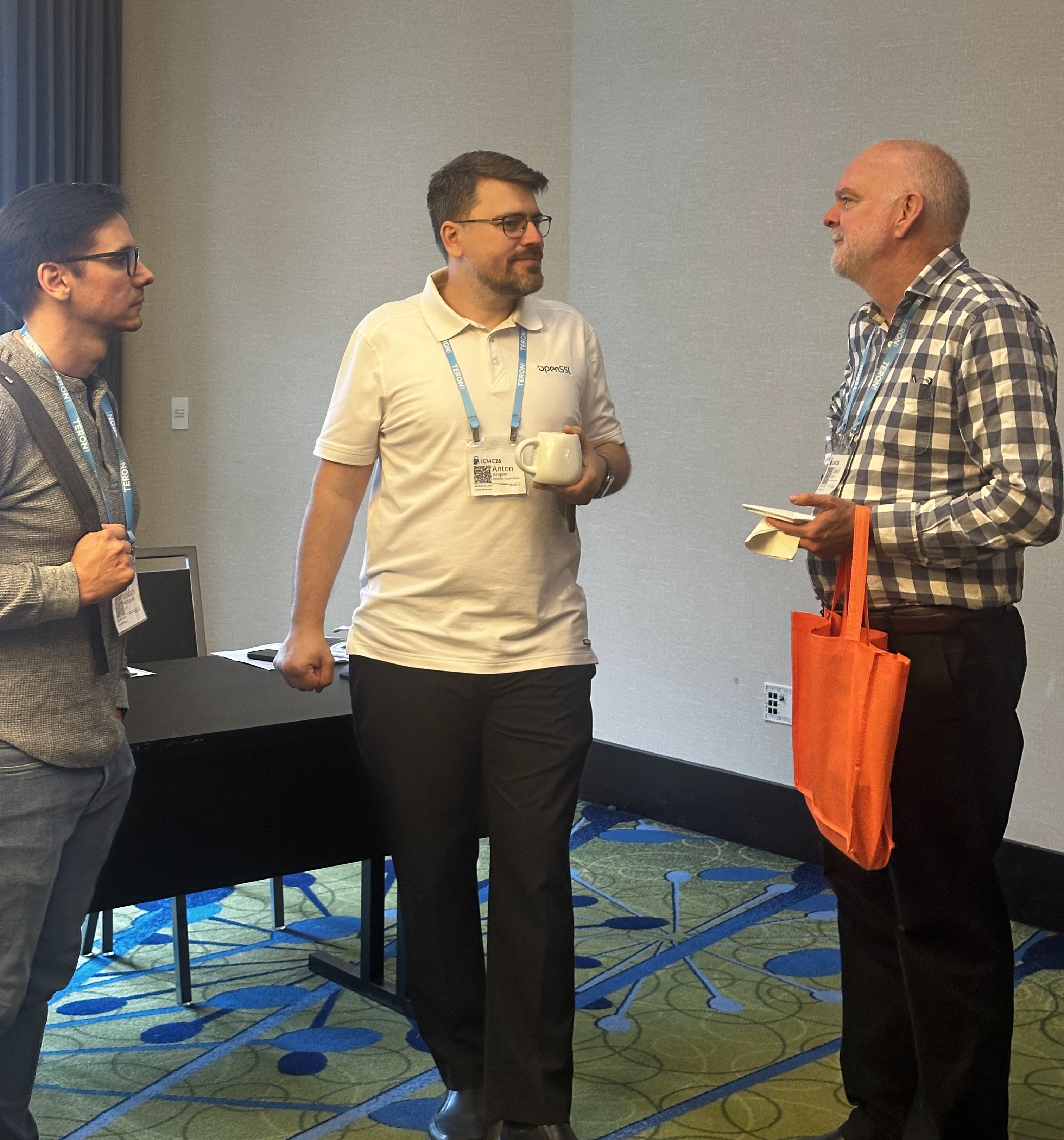 Anton Arapov in an OpenSSL polo holding a coffee mug, speaking with a bearded attendee in a checkered shirt, third person listening on the left.