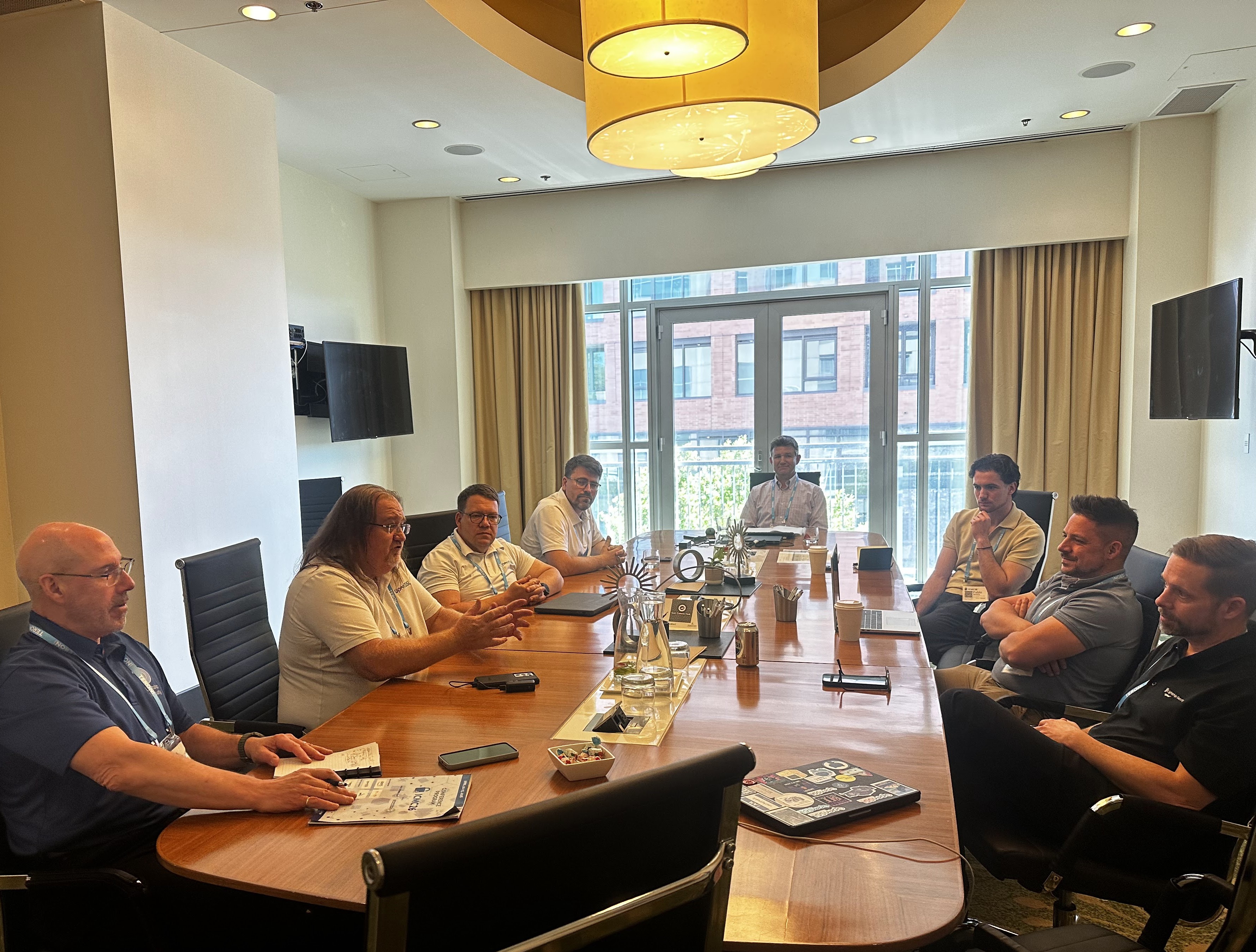 OpenSSL Corporation management team in a boardroom meeting with Lightship FIPS lab, eight people seated around a wood-grain table, natural light from tall windows, James Ramage speaking.