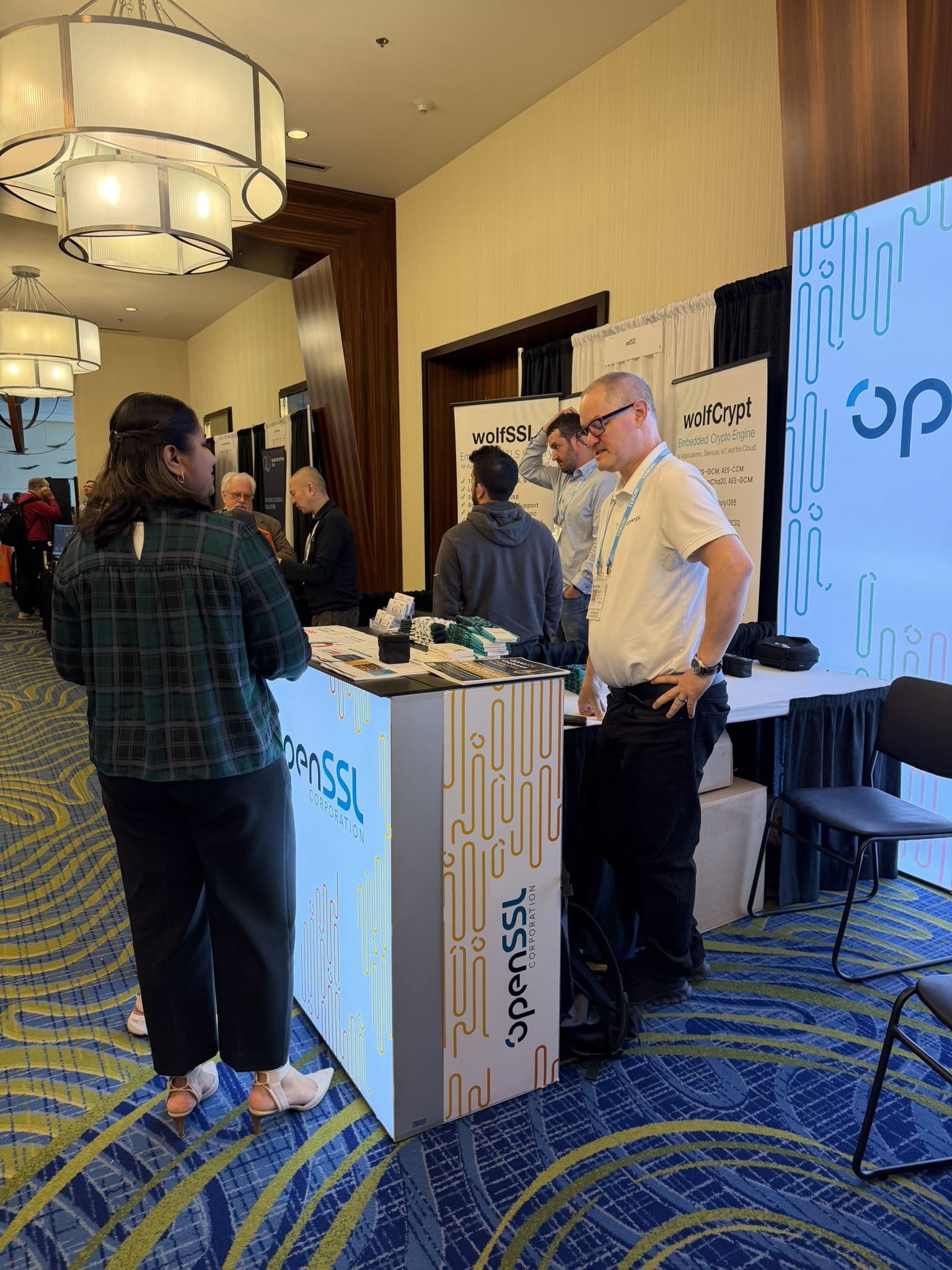 OpenSSL booth at ICMC26, Tim in an OpenSSL polo speaking with a visitor in a plaid shirt, wolfSSL and wolfCrypt banners visible to the right.