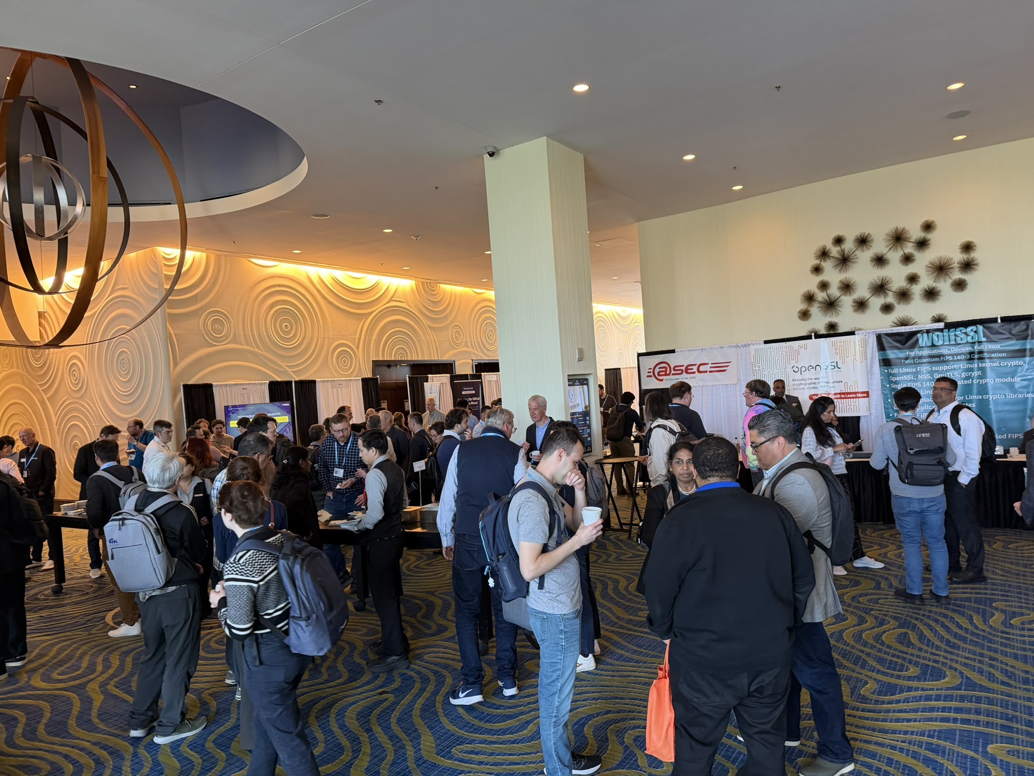 The ICMC26 exhibit corridor during a coffee break — dozens of attendees in clusters, sponsor banners for atsec, OpenSSL and wolfSSL visible on the right, the ring chandelier overhead, patterned carpet underfoot.