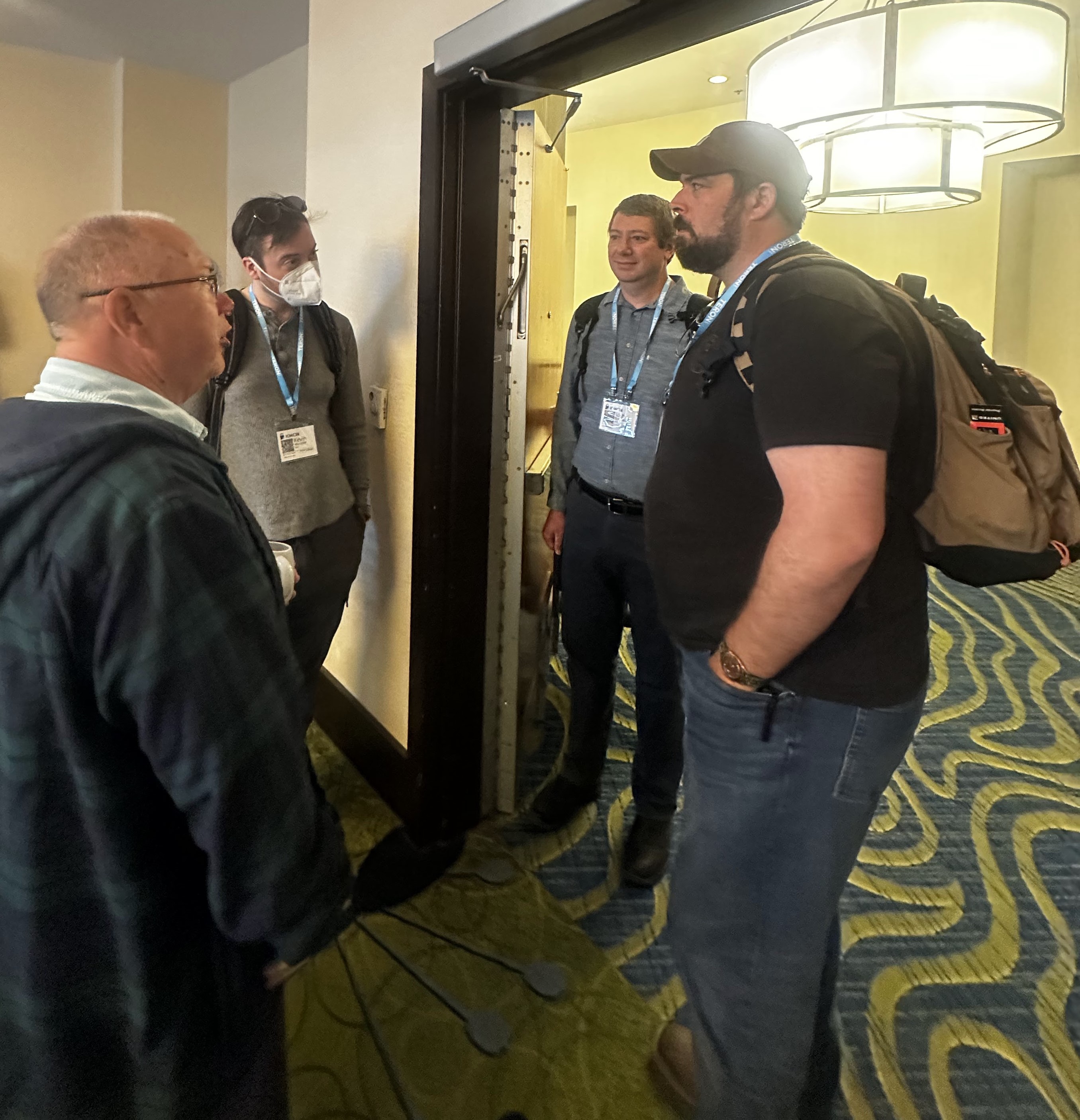 Four attendees in a corridor conversation outside Studio A — one in tartan on the left, one masked in the middle-left, two on the right.