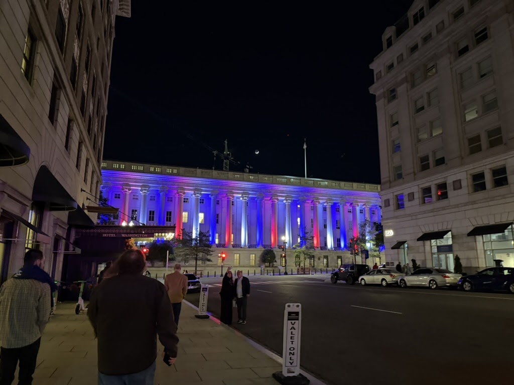 Hotel Washington at night, its colonnade lit in red, white and blue, pedestrians walking toward it.