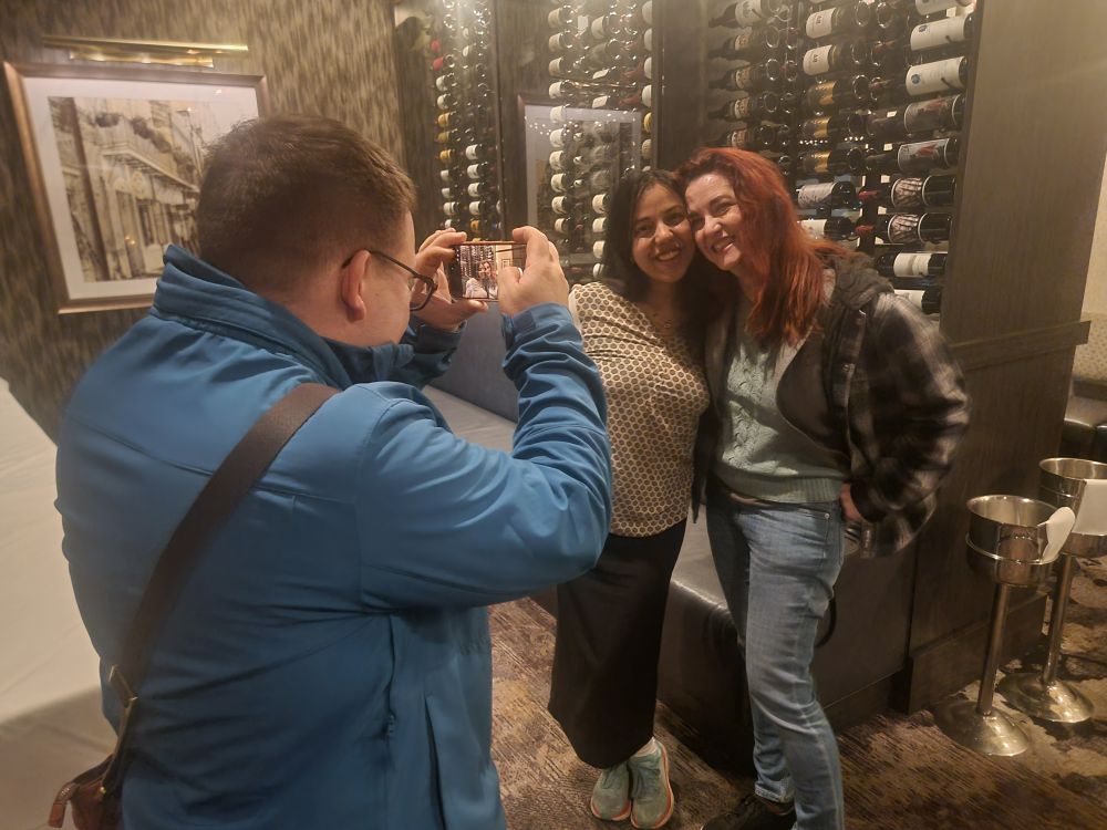 Two attendees posing together in front of a wall of wine bottles, a third attendee photographing them on a phone.