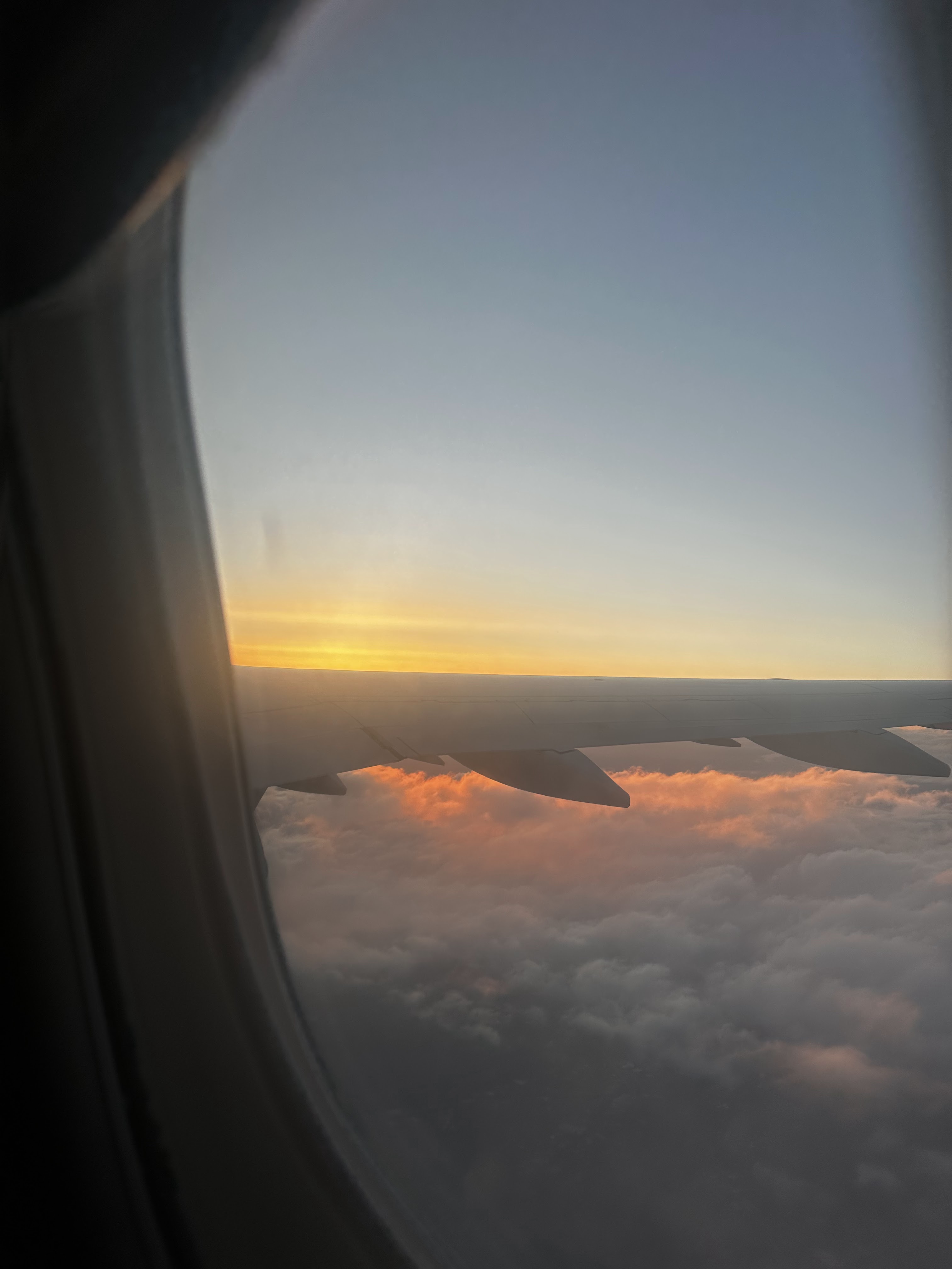 Airplane wing seen through a cabin window, sunrise bleeding orange along the horizon, clouds in soft pink below.